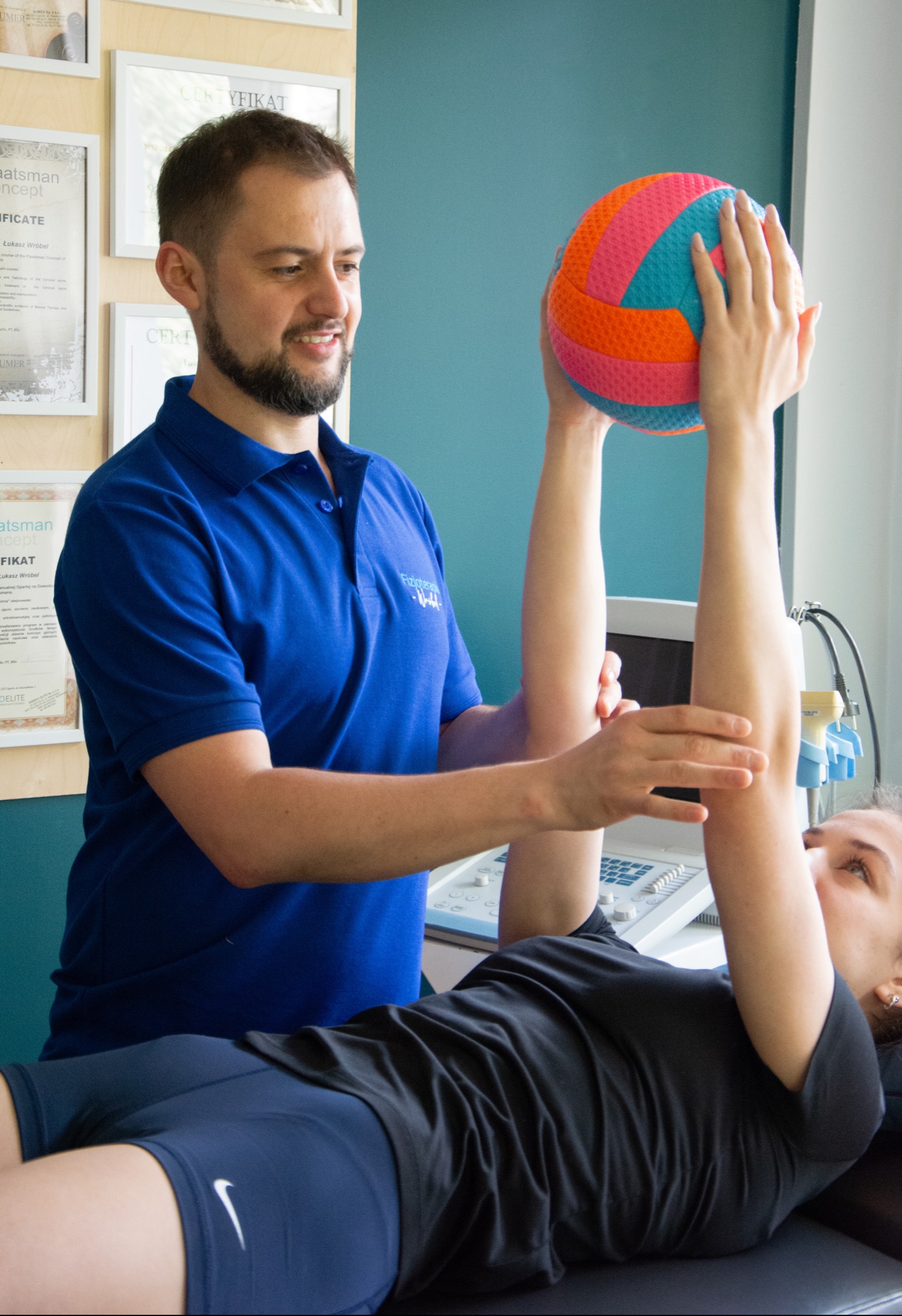 Pacjentka leżąca na plecach z uniesionymi rękami trzyma kolorową piłkę pod nadzorem specjalisty w niebieskiej koszulce, w tle ściana w kolorze turkusowym i certyfikaty na ścianie.