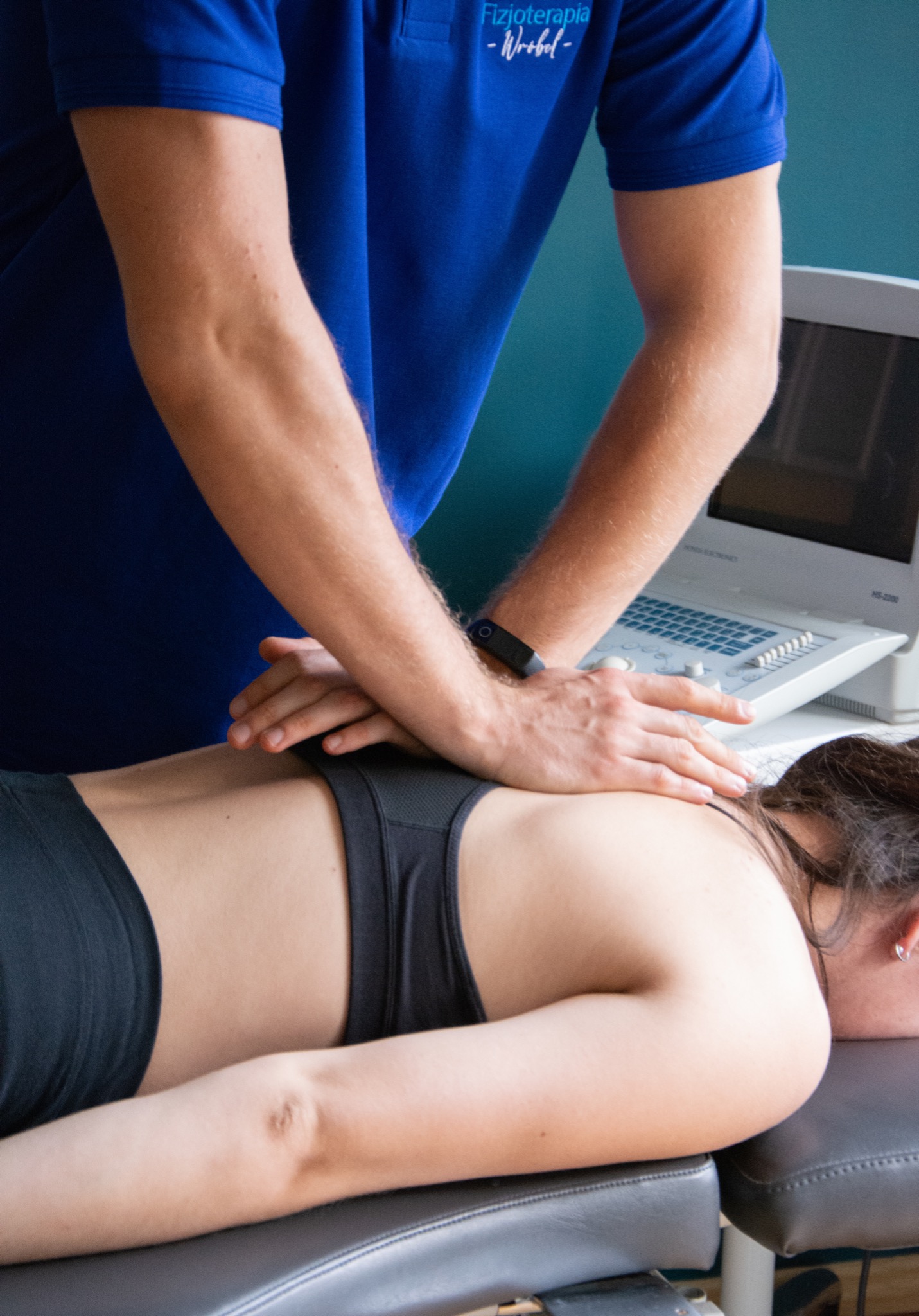Pacjentka leżąca na brzuchu podczas terapii manualnej kręgosłupa, wykonywanej przez terapeutę w niebieskiej koszulce z logo, widoczny aparat USG w tle.