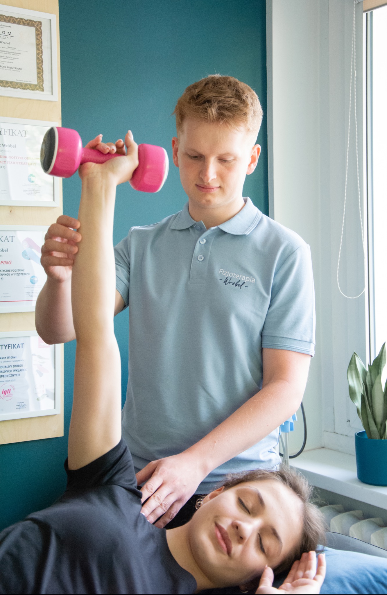 Pacjentka leżąca na plecach, podnosi ramię z różowym hantlem, asystuje jej terapeuta w jasnoniebieskiej koszulce polo z logo, w tle ściana z certyfikatami i roślina doniczkowa.