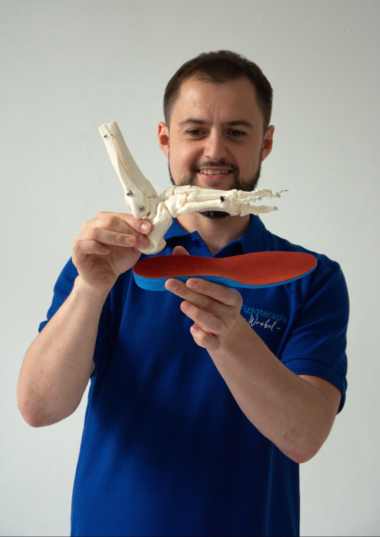 Uśmiechnięty fizjoterapeuta prezentuje model kości stopy oraz wkładkę ortopedyczną z logo firmy 'Fizjoterapia Wróbel'.