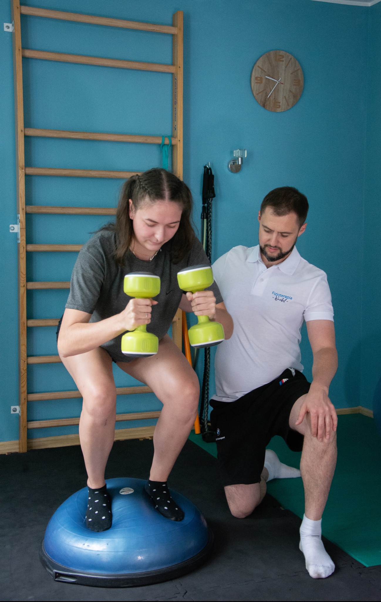 Kobieta w szarym t-shircie i skarpetkach w kropki ćwiczy z zielonymi hantlami, stojąc na niebieskiej platformie BOSU, pod nadzorem mężczyzny w białej koszulce polo z logo na tle drabinki...