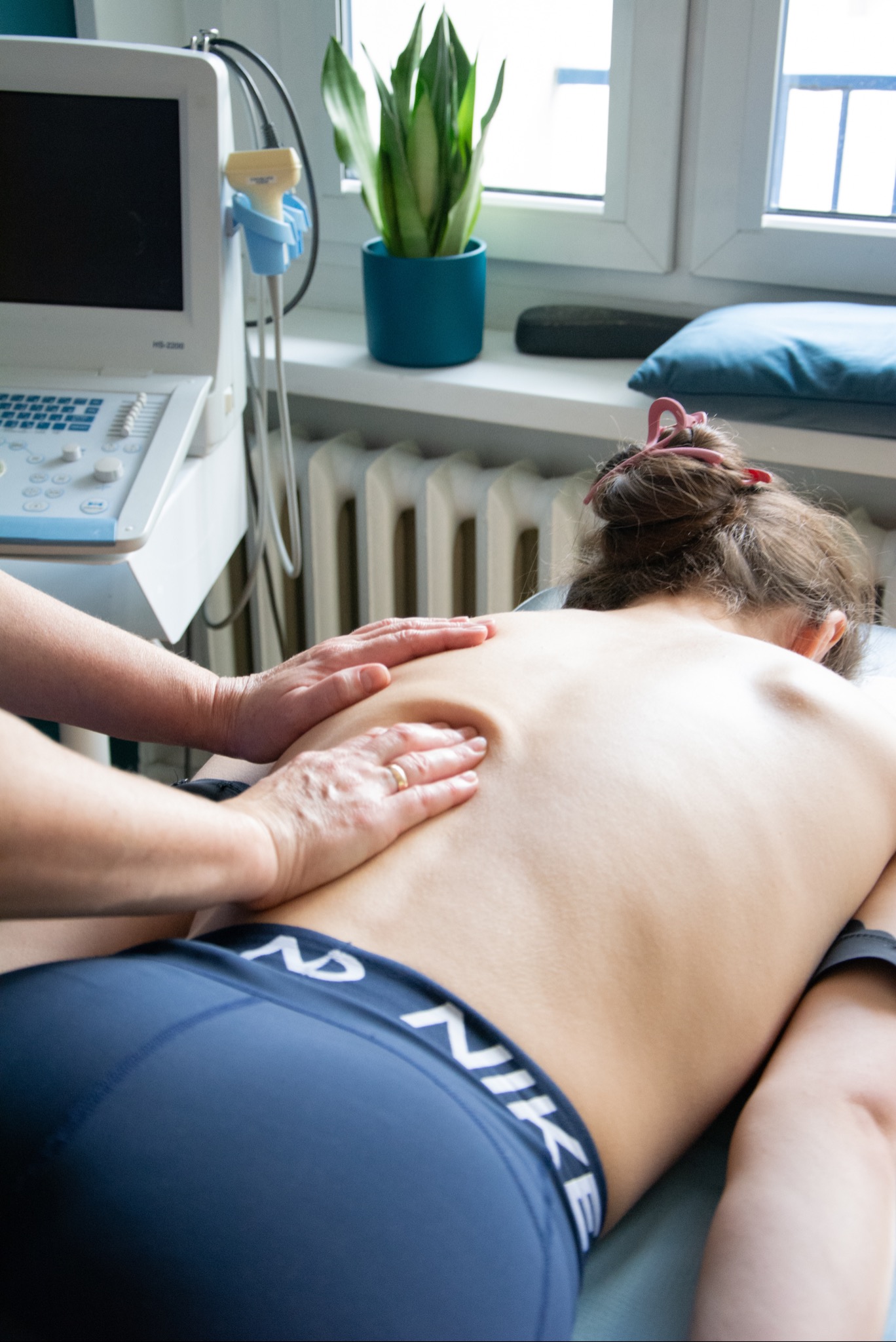 Masaż pleców pacjenta leżącego na brzuchu, wykonywany przez terapeutę dłońmi, w tle aparat USG i roślina doniczkowa przy oknie.