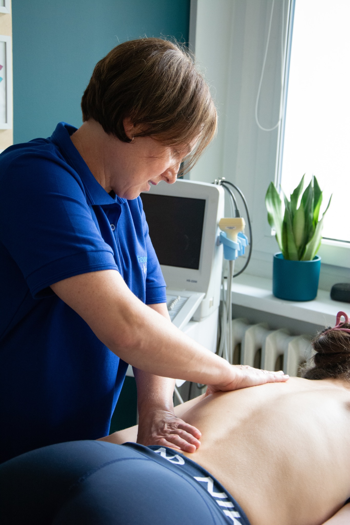 Kobieta w niebieskiej koszulce z logo wykonuje masaż pleców pacjentowi w gabinecie z aparaturą do fizykoterapii i rośliną doniczkową na parapecie.