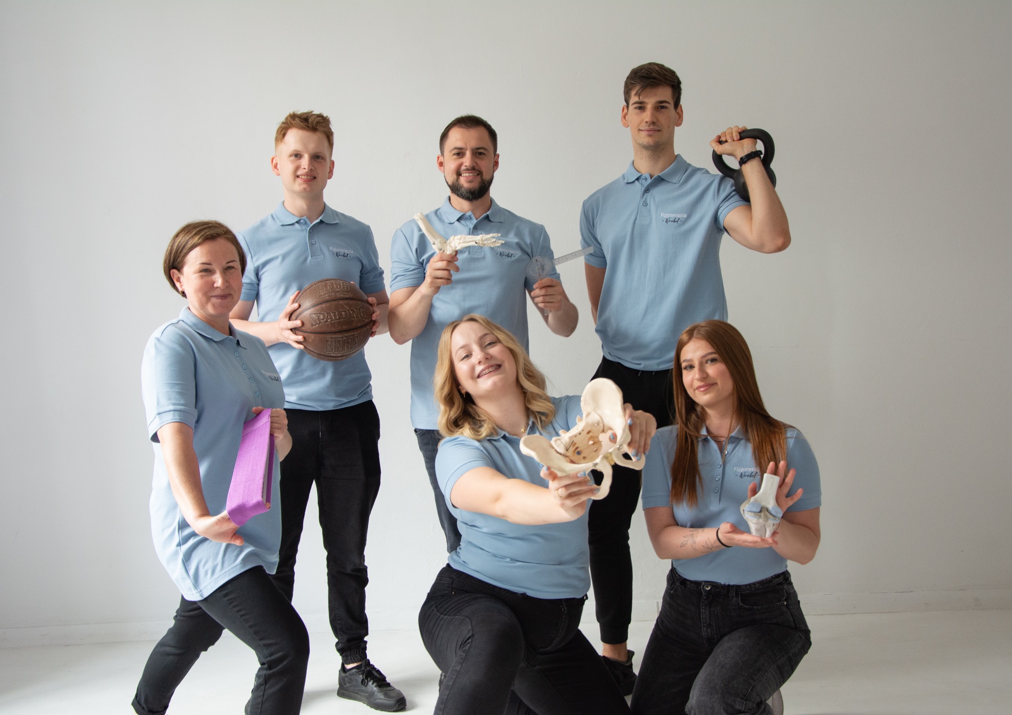 Grupa sześciu osób w błękitnych koszulkach polo prezentuje modele anatomiczne kości, piłkę do koszykówki, kettlebell i taśmę do ćwiczeń na tle białej ściany. Na koszulkach widoczne logo firmy.