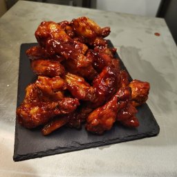 Kuchnia nr 13, LECH URBANOWSKI - Góra skrzydełek kurczaka w słodko-pikantnym sosie barbecue, ułożona na czarnym, kamiennym talerzu, widoczna na metalowym blacie kuchennym w profesjonalnej kuchni.