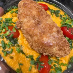 Gotowe danie w plastikowym opakowaniu: panierowany filet rybny na pomarańczowym puree, udekorowany pomidorkami cherry i posiekanym szczypiorkiem.