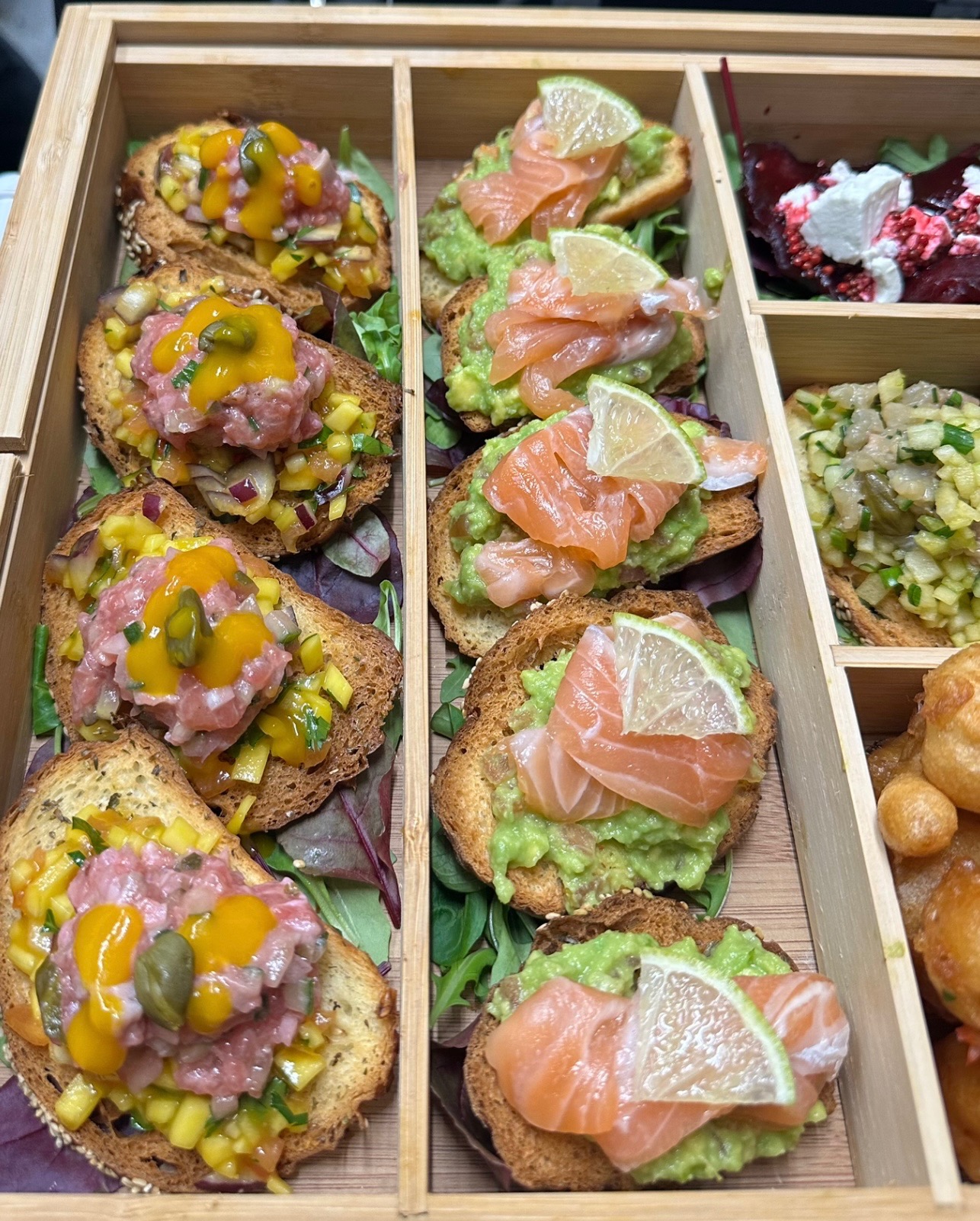 Drewniana taca podzielona na przegródki prezentująca różnorodne przekąski: grzanki z tatarem i mango, grzanki z awokado i łososiem z plasterkiem limonki, sałatka z buraków z serem oraz kompozycja...