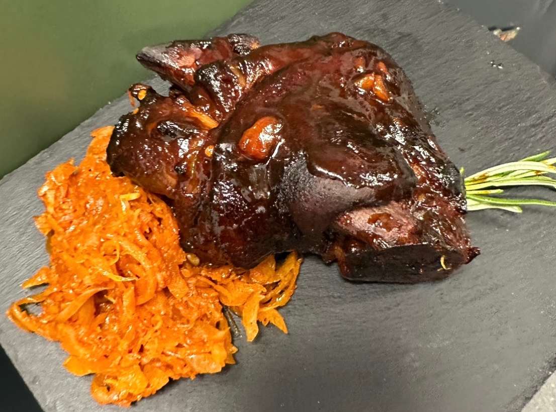 Glazurowana golonka wieprzowa z dodatkiem duszonej marchewki i gałązką rozmarynu, podana na czarnym, kamiennym talerzu.