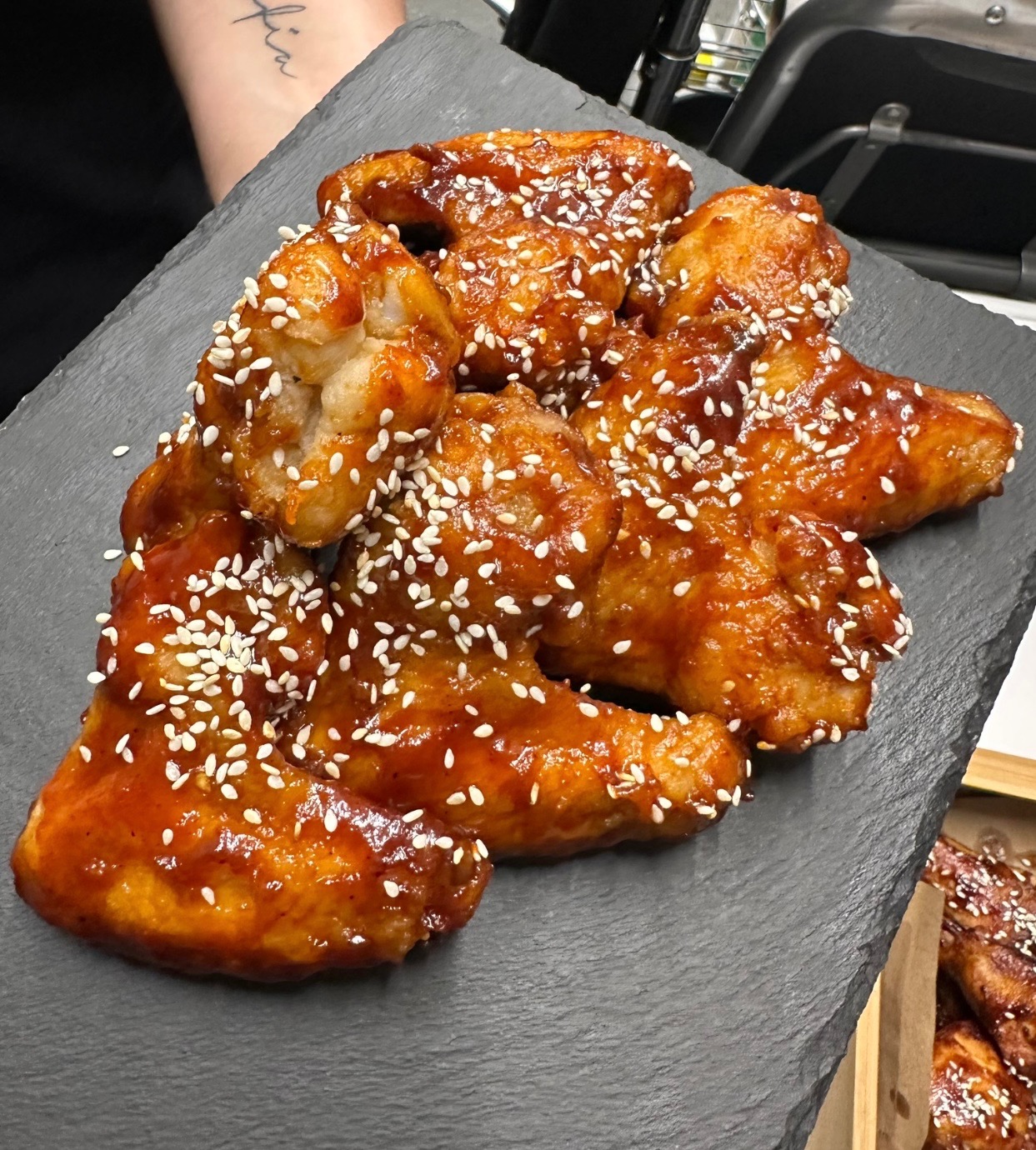 Glazurowane skrzydełka kurczaka posypane sezamem, ułożone na czarnym, łupkowym talerzu, z widocznym fragmentem tatuażu na ręce osoby trzymającej talerz.