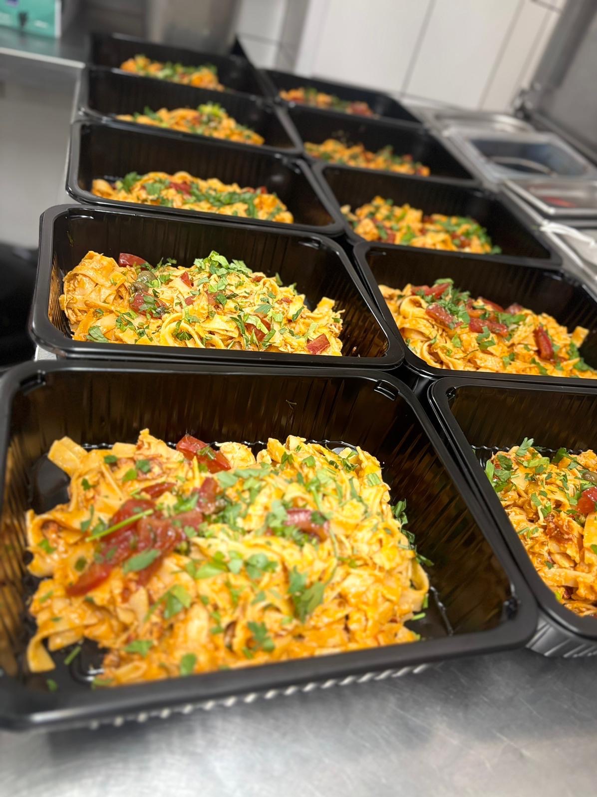 Rząd czarnych pojemników z makaronem tagliatelle w sosie pomidorowym, posypanym świeżą pietruszką i kawałkami suszonych pomidorów, gotowe do transportu.