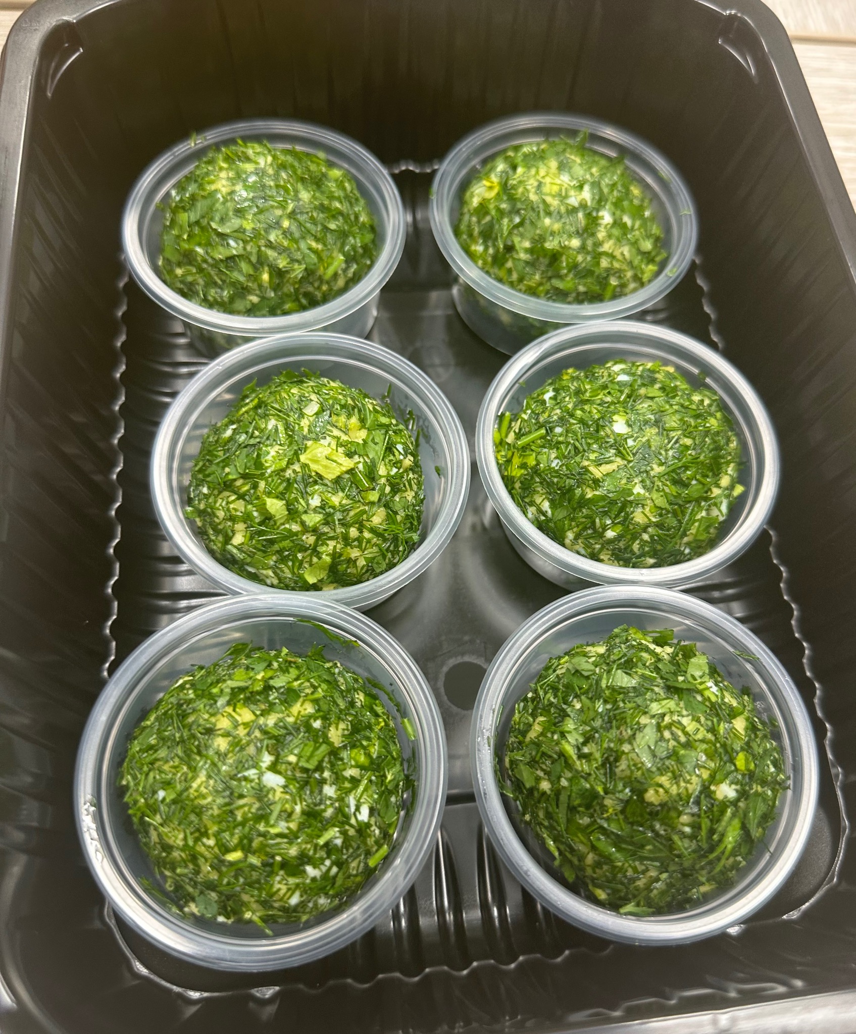 Sześć porcji kul serowych obtoczonych posiekanymi ziołami w plastikowych pojemnikach, ułożonych w czarnym opakowaniu zbiorczym.