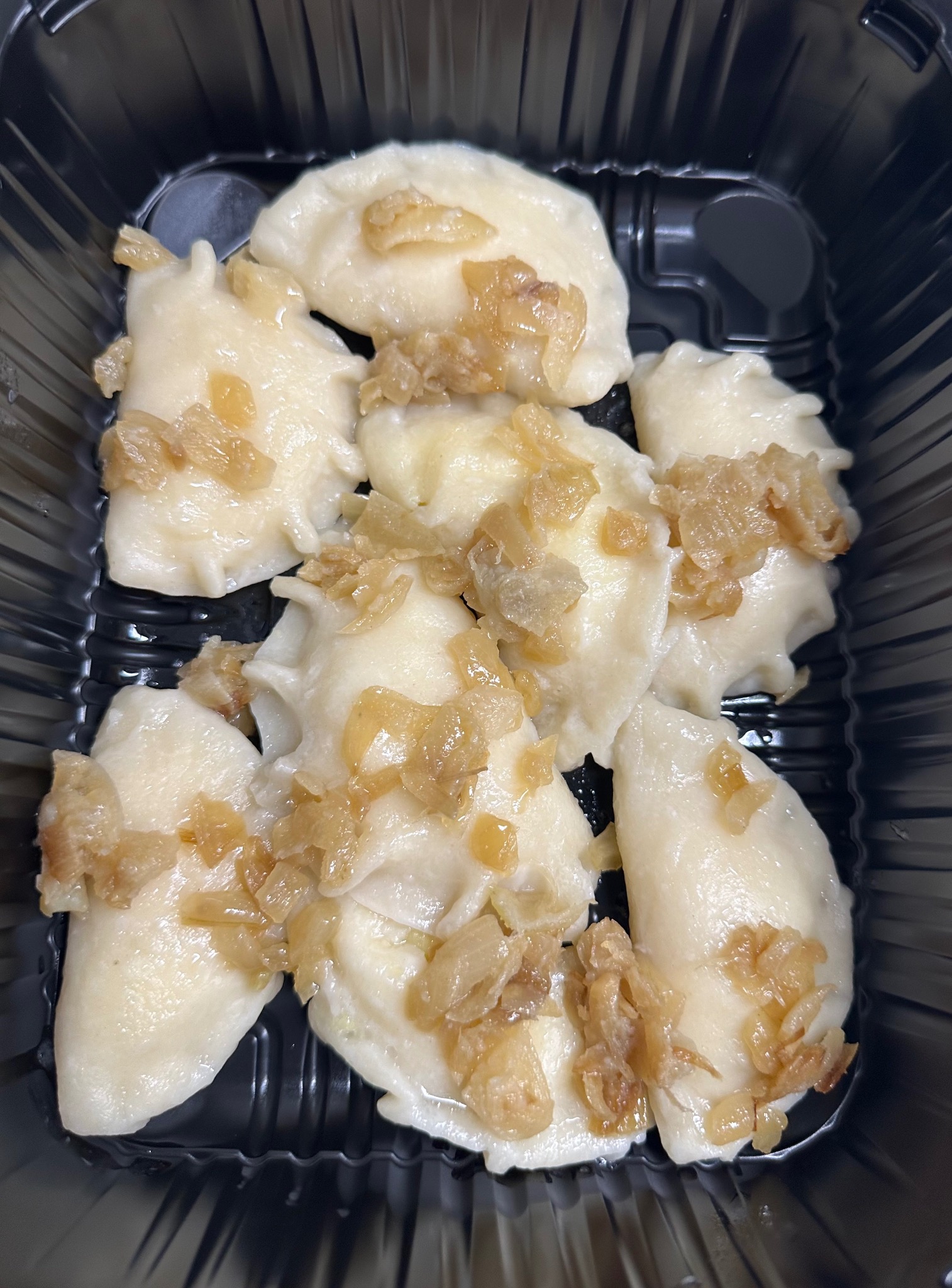 Porcja gotowych pierogów z cebulką, ułożonych w czarnym, plastikowym pojemniku na wynos.