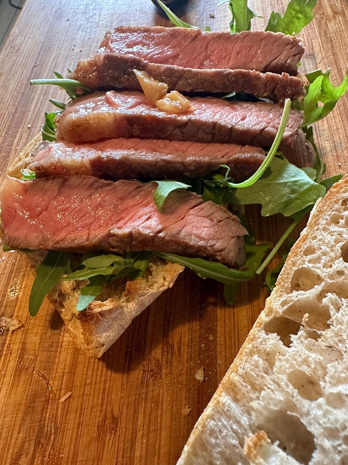 Kanapka z rostbefem i rukolą na drewnianej desce, z widocznym kawałkiem chleba obok.