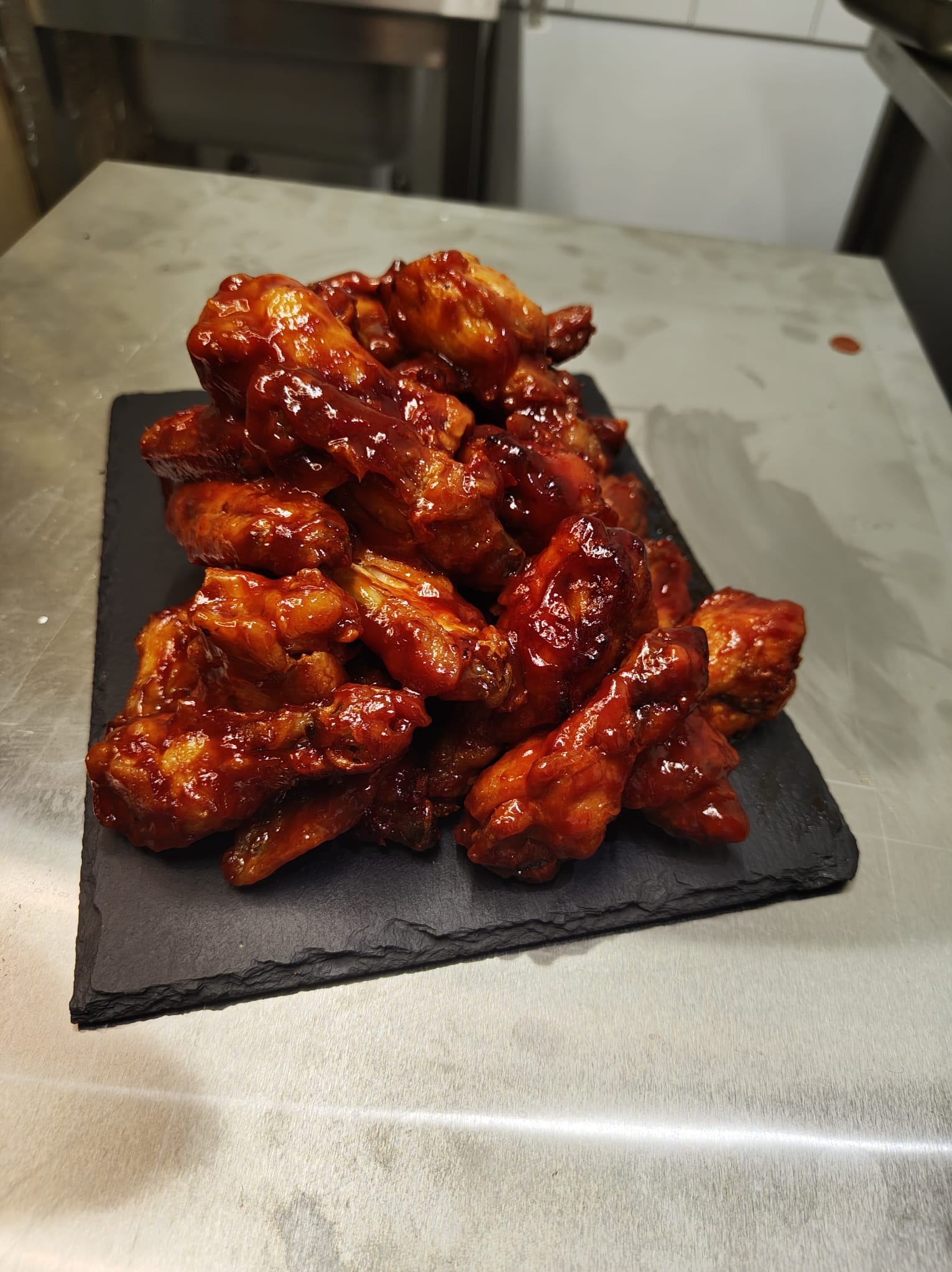 Góra skrzydełek kurczaka w słodko-pikantnym sosie barbecue, ułożona na czarnym, kamiennym talerzu, widoczna na metalowym blacie kuchennym w profesjonalnej kuchni.