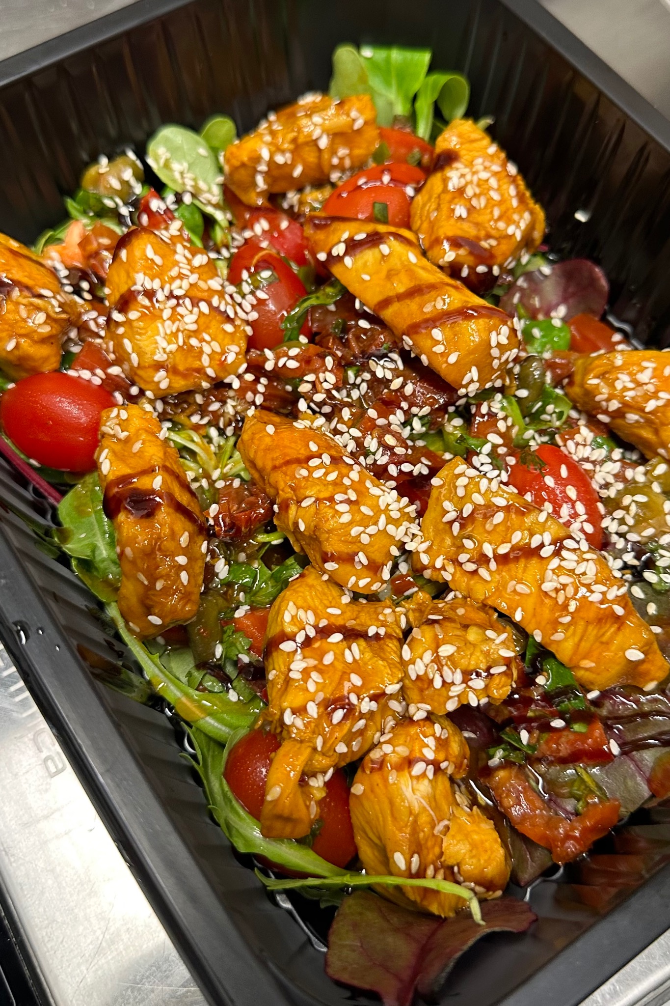 Sałatka z grillowanym kurczakiem w sosie teriyaki, posypana sezamem, podana w czarnym, prostokątnym pojemniku na tle blatu ze stali nierdzewnej.