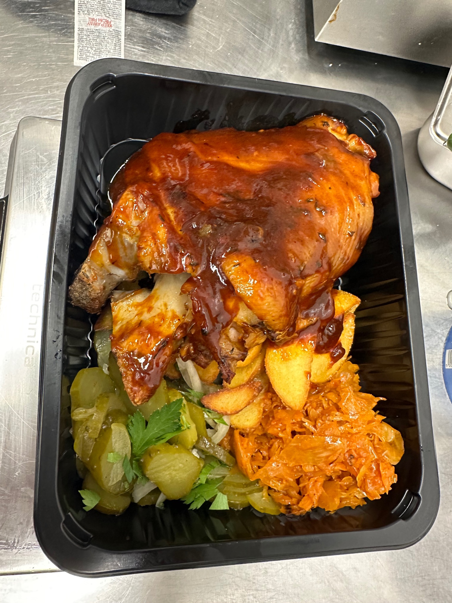 Porcja obiadowa w czarnym opakowaniu: pieczone udo kurczaka polane sosem BBQ, ziemniaki pieczone w łupinach, kapusta duszona i sałatka z ogórków konserwowych z natką pietruszki.