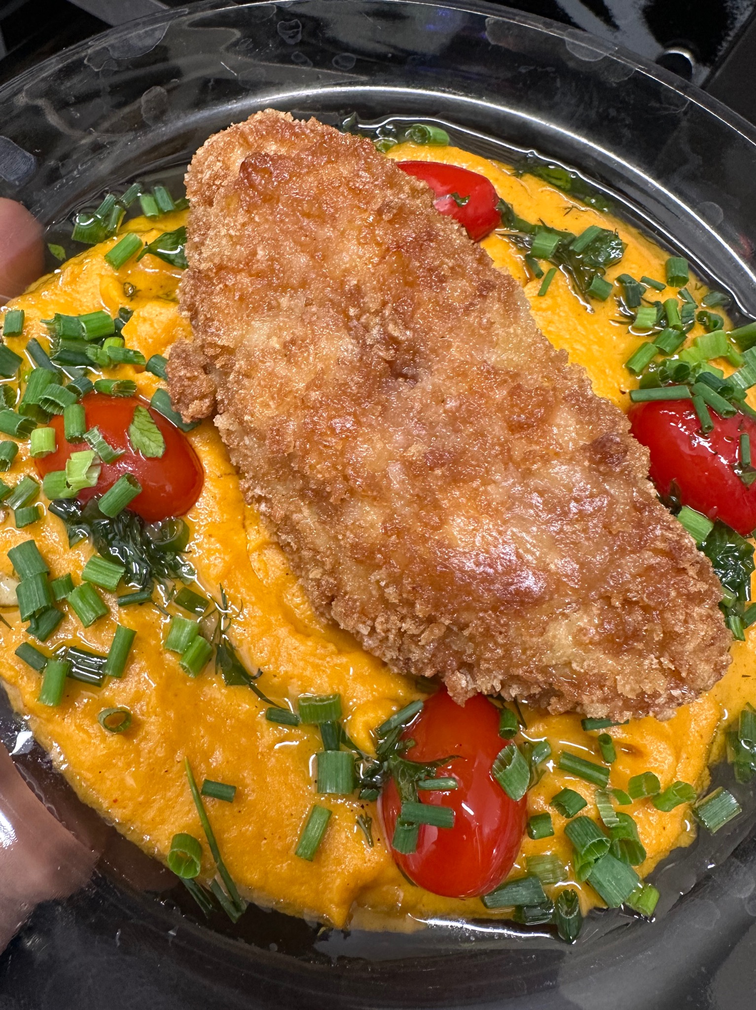 Gotowe danie w plastikowym opakowaniu: panierowany filet rybny na pomarańczowym puree, udekorowany pomidorkami cherry i posiekanym szczypiorkiem.