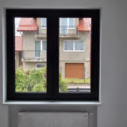 Dwuskrzydłowe okno w kolorze antracytowym, z widokiem na budynek mieszkalny z czerwoną dachówką i zielonymi krzewami, zamontowane w jasnym pomieszczeniu z grzejnikiem pod parapetem.