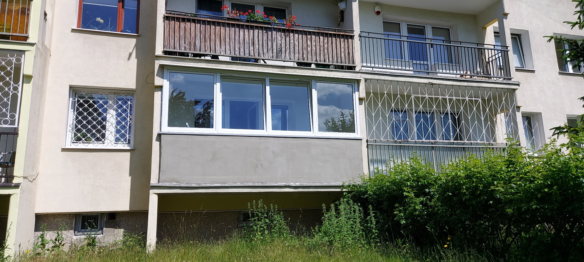 Fasada bloku z balkonami, na jednym z balkonów zamontowane białe okna, na pozostałych balkonach balustrady. W oknach widoczne odbicie nieba.