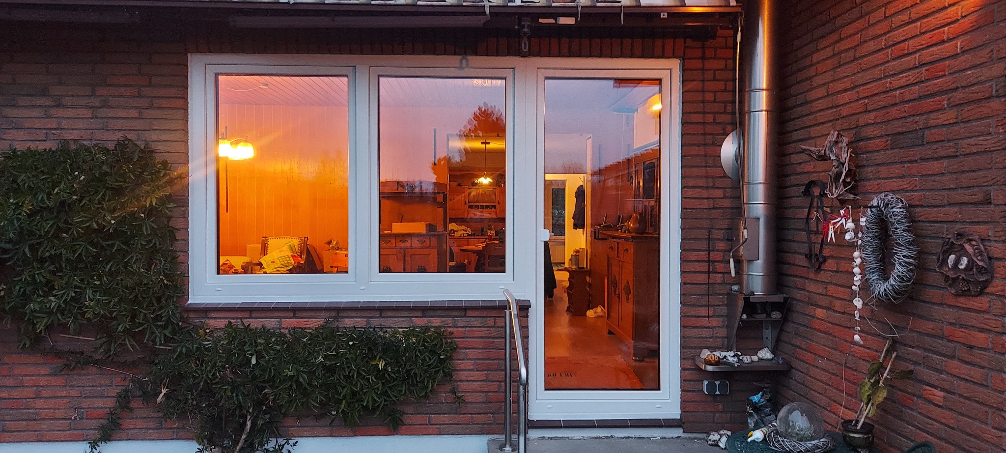 Białe okno z drzwiami balkonowymi na tle ceglanej ściany domu, z widocznym wnętrzem o ciepłym oświetleniu i dekoracjami na ścianie.