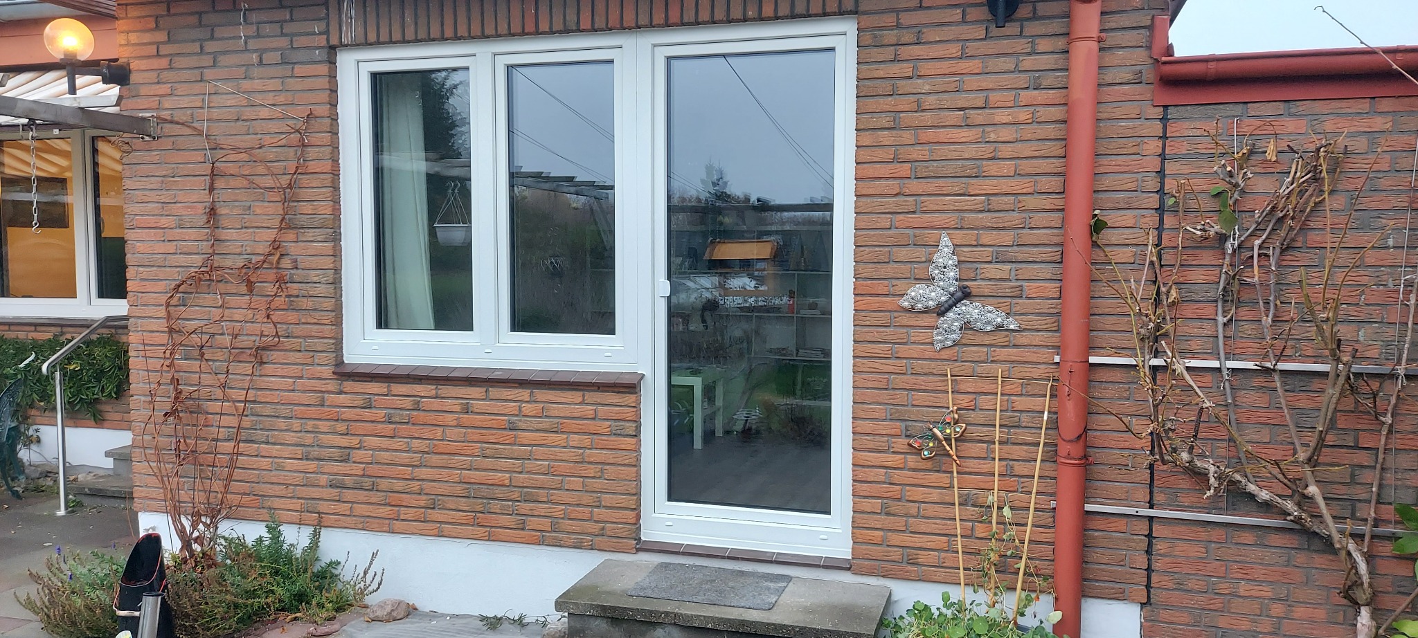 Białe okno PCV z drzwiami balkonowymi w ceglanej ścianie domu, widoczne dekoracje ścienne w postaci motyla i ważki.