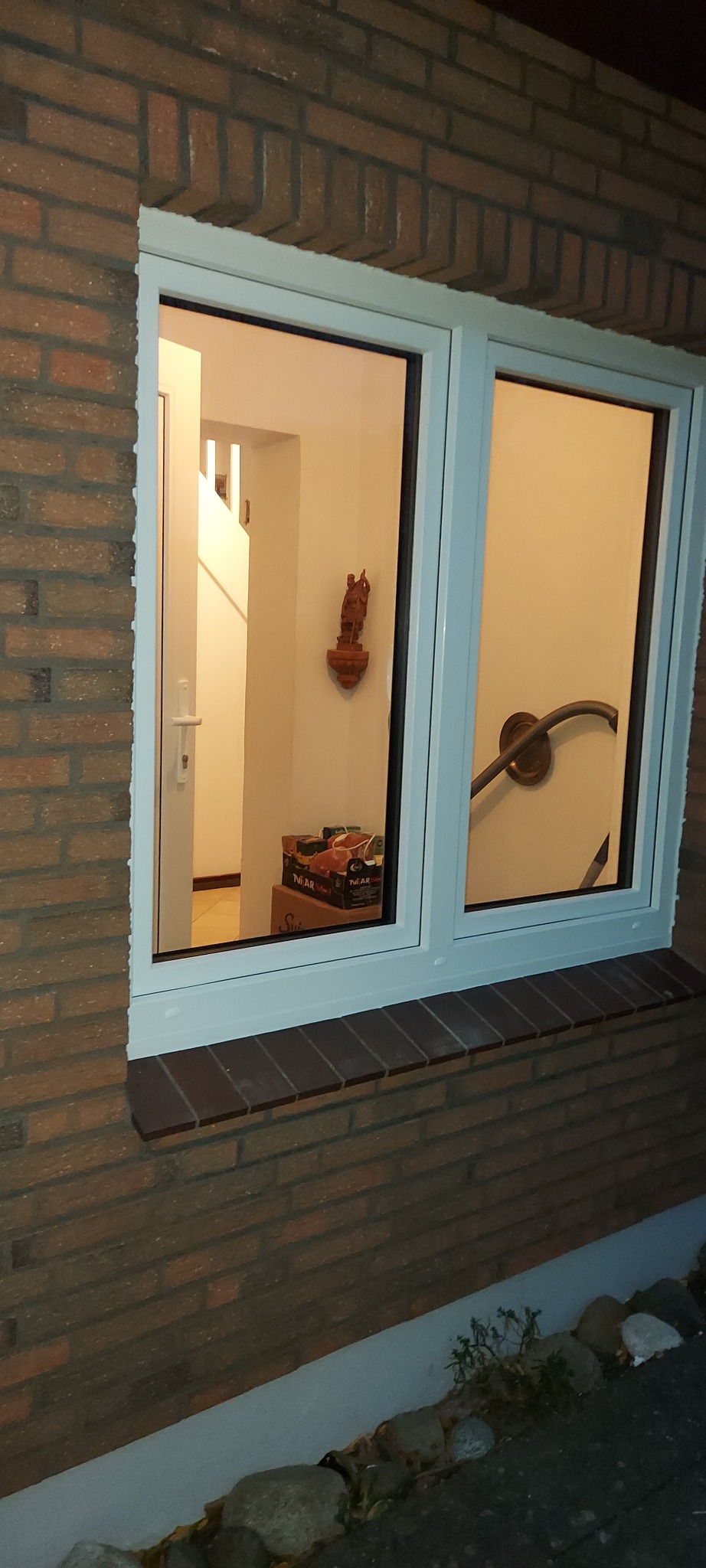 Białe, dwuskrzydłowe okno w murze z cegły. W tle widoczne schody, drzwi oraz dekoracyjna figurka na ścianie.