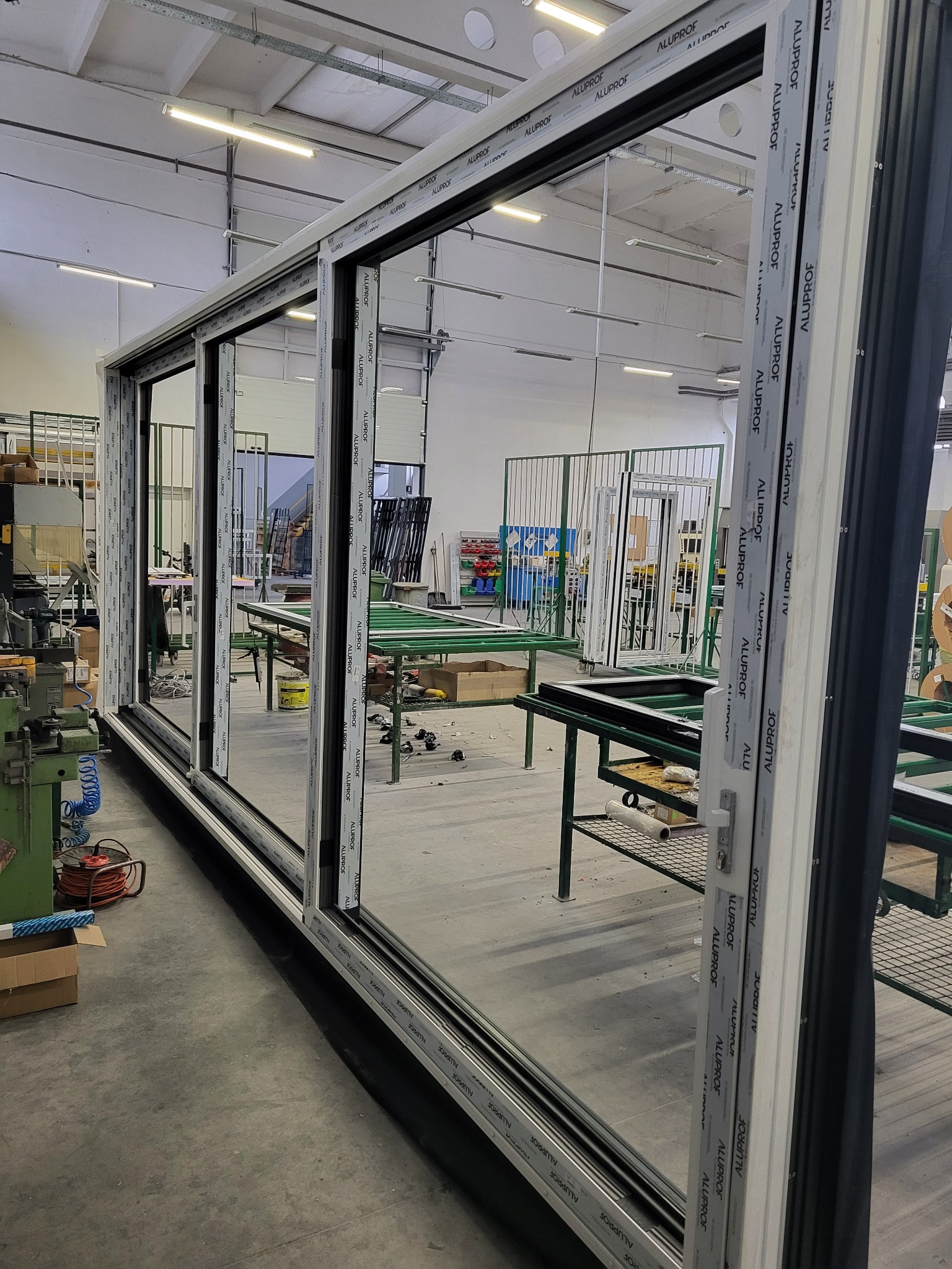 Wielkoformatowe okno przesuwne z profili aluminiowych w hali produkcyjnej, widoczne detale konstrukcyjne i logo producenta na profilach.