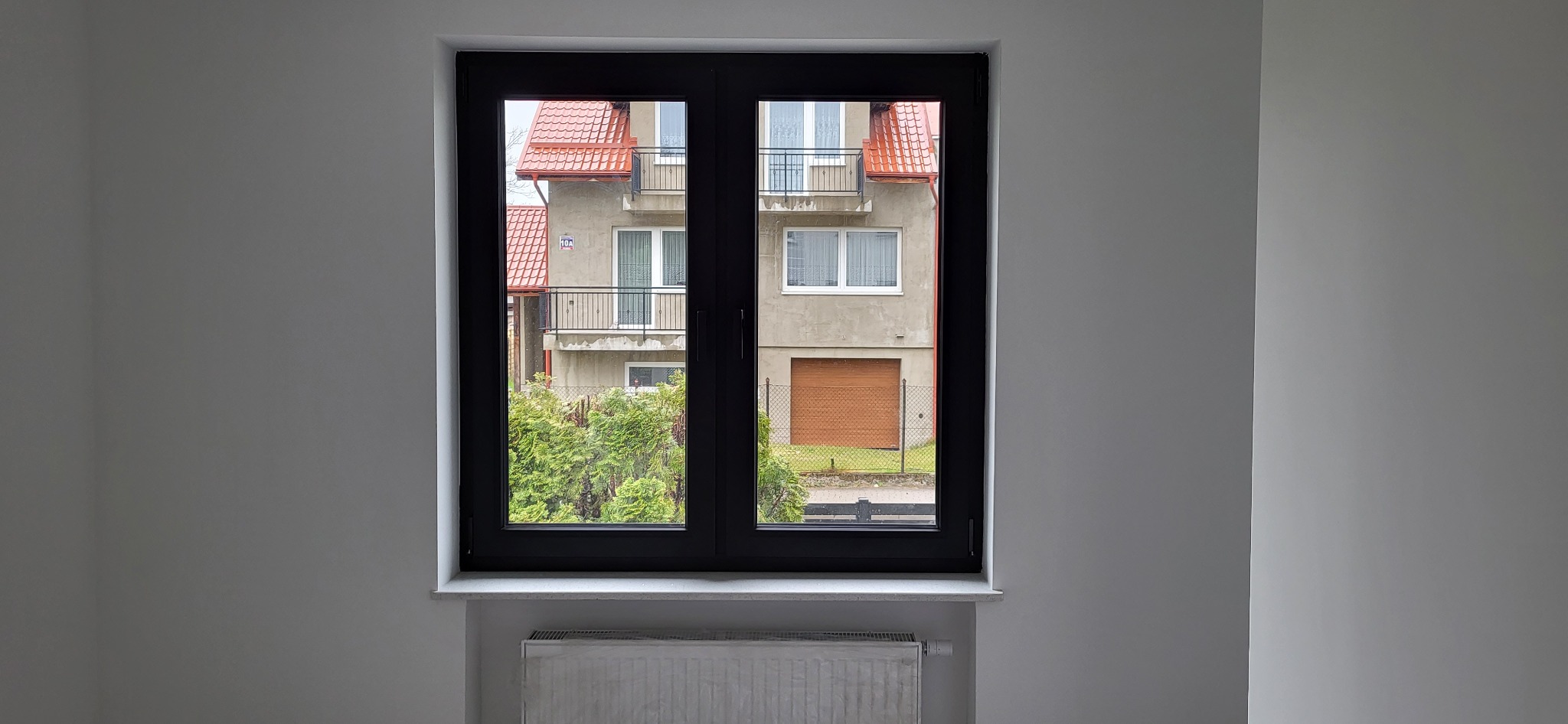 Dwuskrzydłowe okno w kolorze antracytowym, z widokiem na budynek mieszkalny z czerwoną dachówką i zielonymi krzewami, zamontowane w jasnym pomieszczeniu z grzejnikiem pod parapetem.