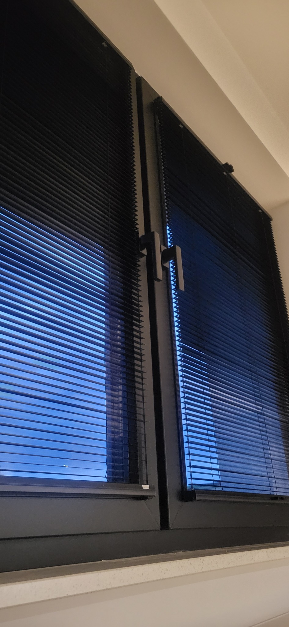 Zamknięte okno PCV z czarnymi żaluzjami w pomieszczeniu z jasnymi ścianami, widok na niebieskie światło dzienne przez lamele.
