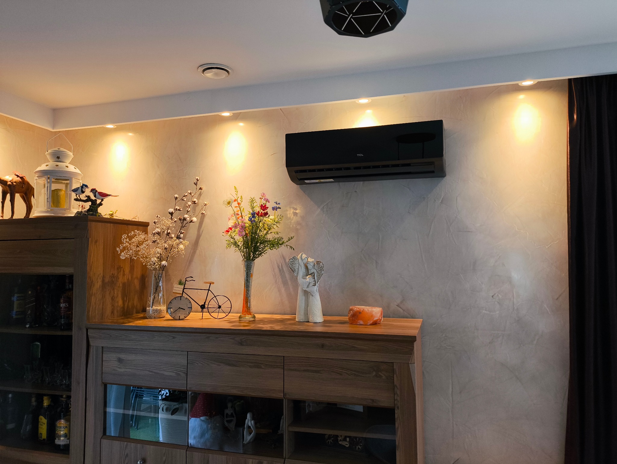 Czarny klimatyzator TCL zamontowany na ścianie w salonie, nad drewnianą komodą z dekoracjami i oświetleniem punktowym.