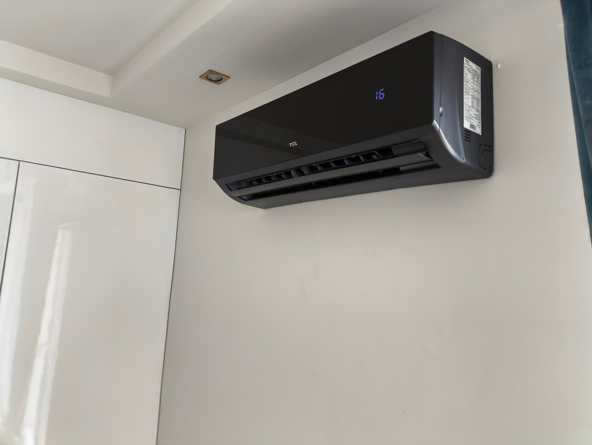 Czarny klimatyzator TCL zamontowany na białej ścianie w pomieszczeniu z białą szafą i oświetleniem punktowym
