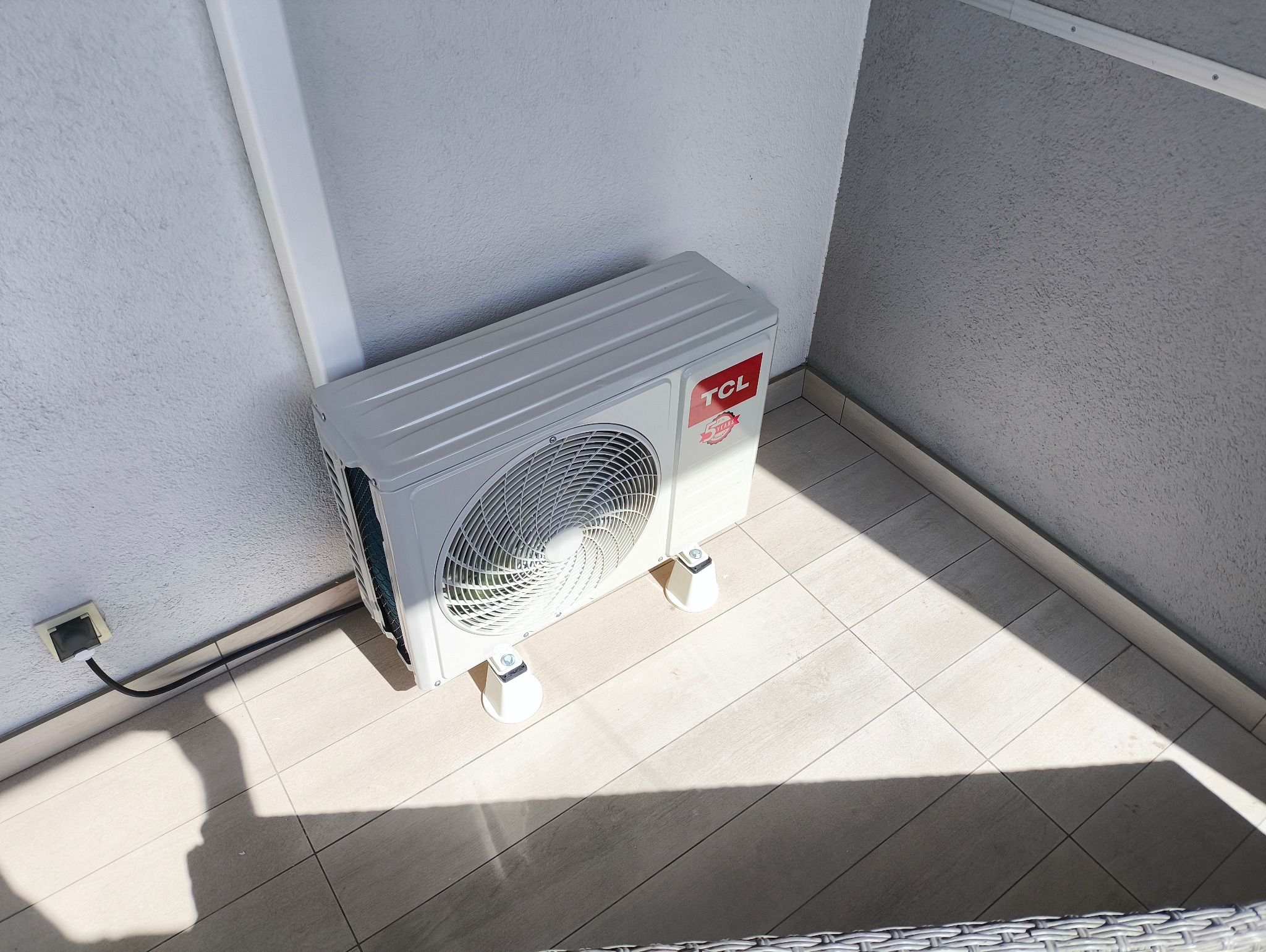 Biała jednostka zewnętrzna klimatyzatora TCL zamontowana na balkonie, widoczne podłączenie elektryczne i cień padający na podłogę z płytek.