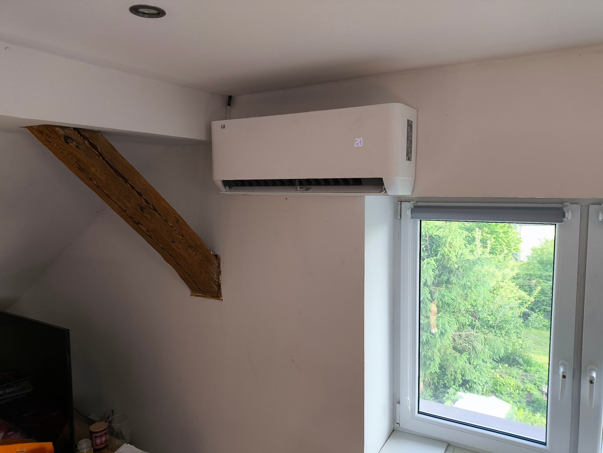 Biała klimatyzacja ścienna zamontowana w pokoju na poddaszu, obok okna i drewnianej belki konstrukcyjnej.