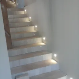 Wykończone marmurowe schody z zamontowanym oświetleniem punktowym LED w ścianie, widoczny fragment listwy przypodłogowej i fragment materiału zabezpieczającego na górze schodów.