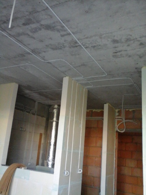 Instalacja elektryczna w surowym wnętrzu: białe przewody prowadzone po betonowym suficie i wzdłuż ścian działowych z cegły, widoczny pion wentylacyjny w izolacji.