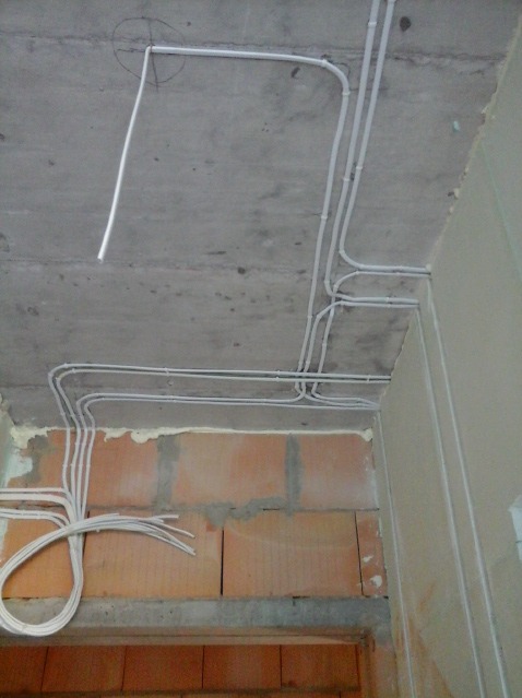 Białe rurki instalacji elektrycznej prowadzone po surowym, betonowym stropie i ceglanej ścianie w trakcie remontu, z wystającymi przewodami w rogu pomieszczenia.