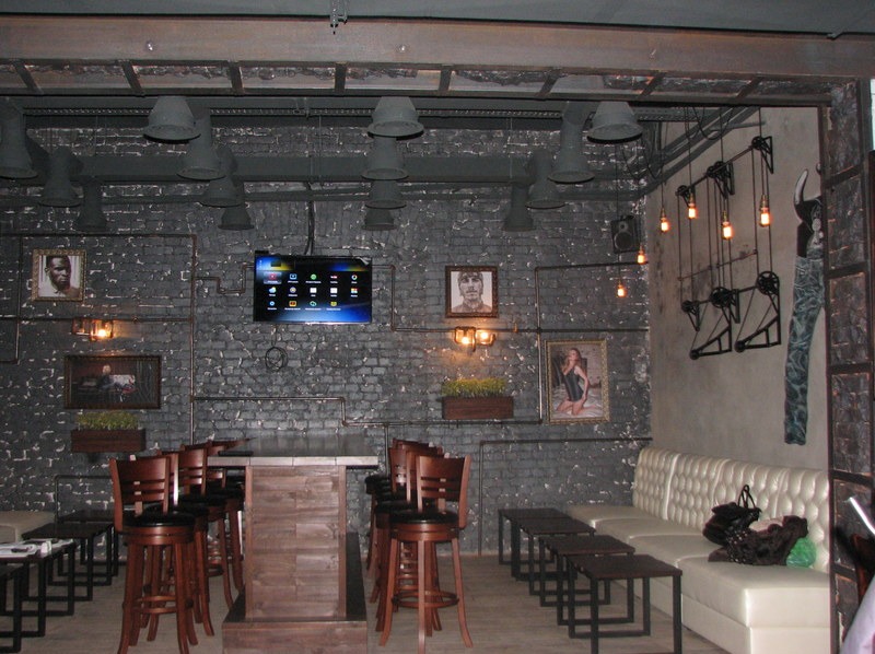 Industrialny bar z ciemną cegłą, widoczne rury instalacji elektrycznej na ścianie, telewizor z menu, oświetlenie w stylu loft i drewniane meble.