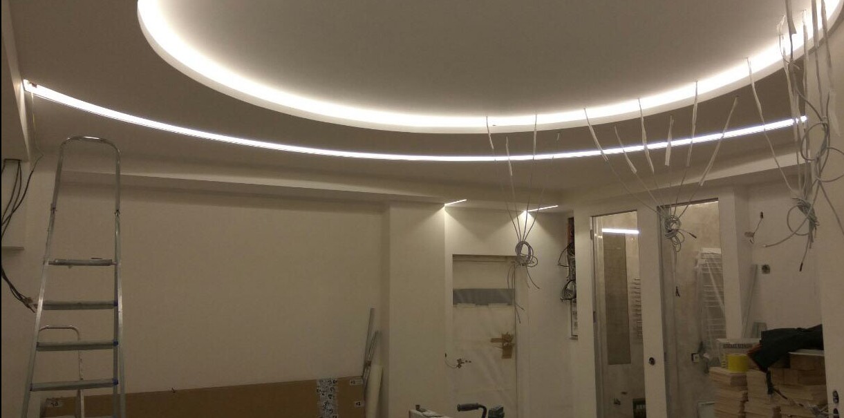 Wnętrze w trakcie montażu oświetlenia sufitowego LED z widocznymi przewodami, drabina aluminiowa oparta o ścianę, elementy wykończeniowe w tle.