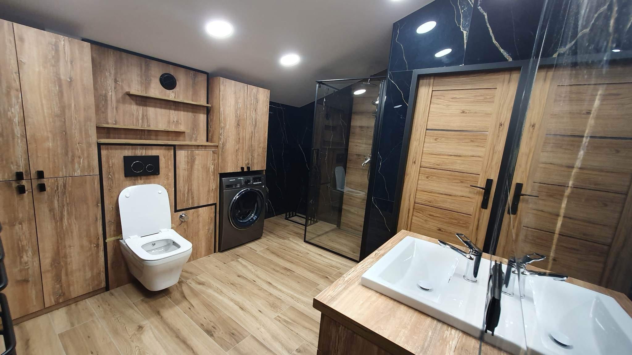 Nowoczesna łazienka z drewnianymi szafkami, czarnym marmurem, kabiną prysznicową ze szklanymi drzwiami, pralką i podwójną umywalką.