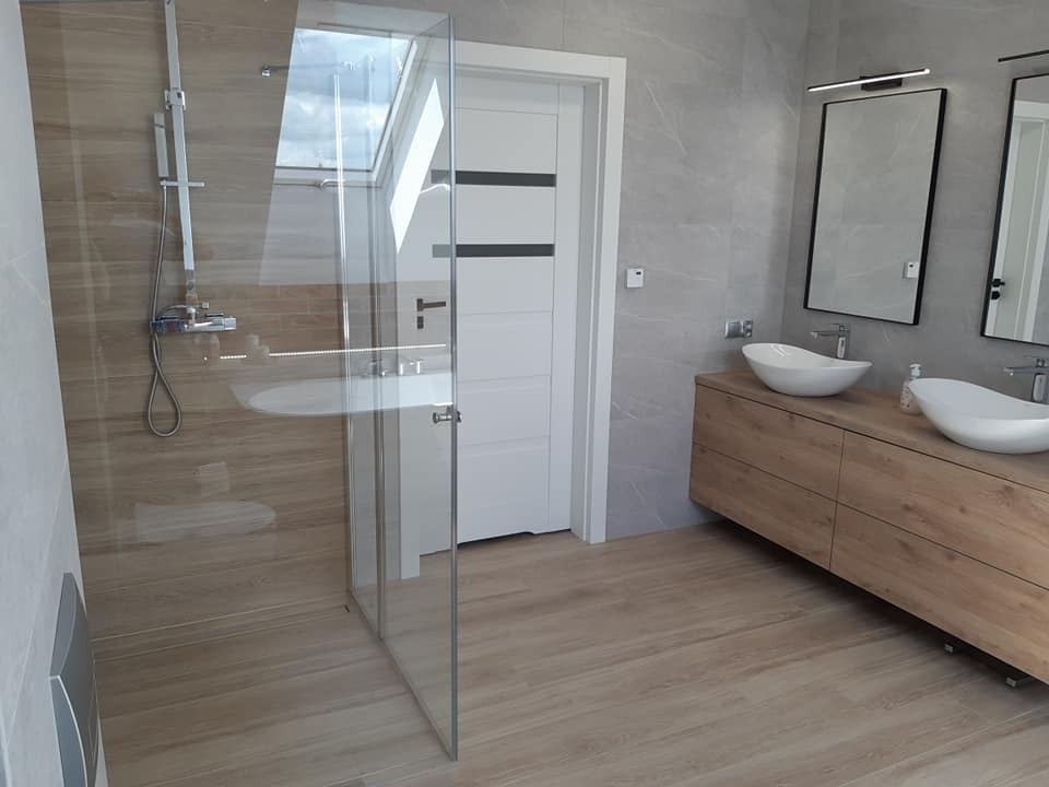 Nowoczesna łazienka z kabiną prysznicową ze szklanymi ściankami, podwójną umywalką nablatową na drewnianej szafce, dużymi lustrami w czarnych ramach i oknem dachowym.