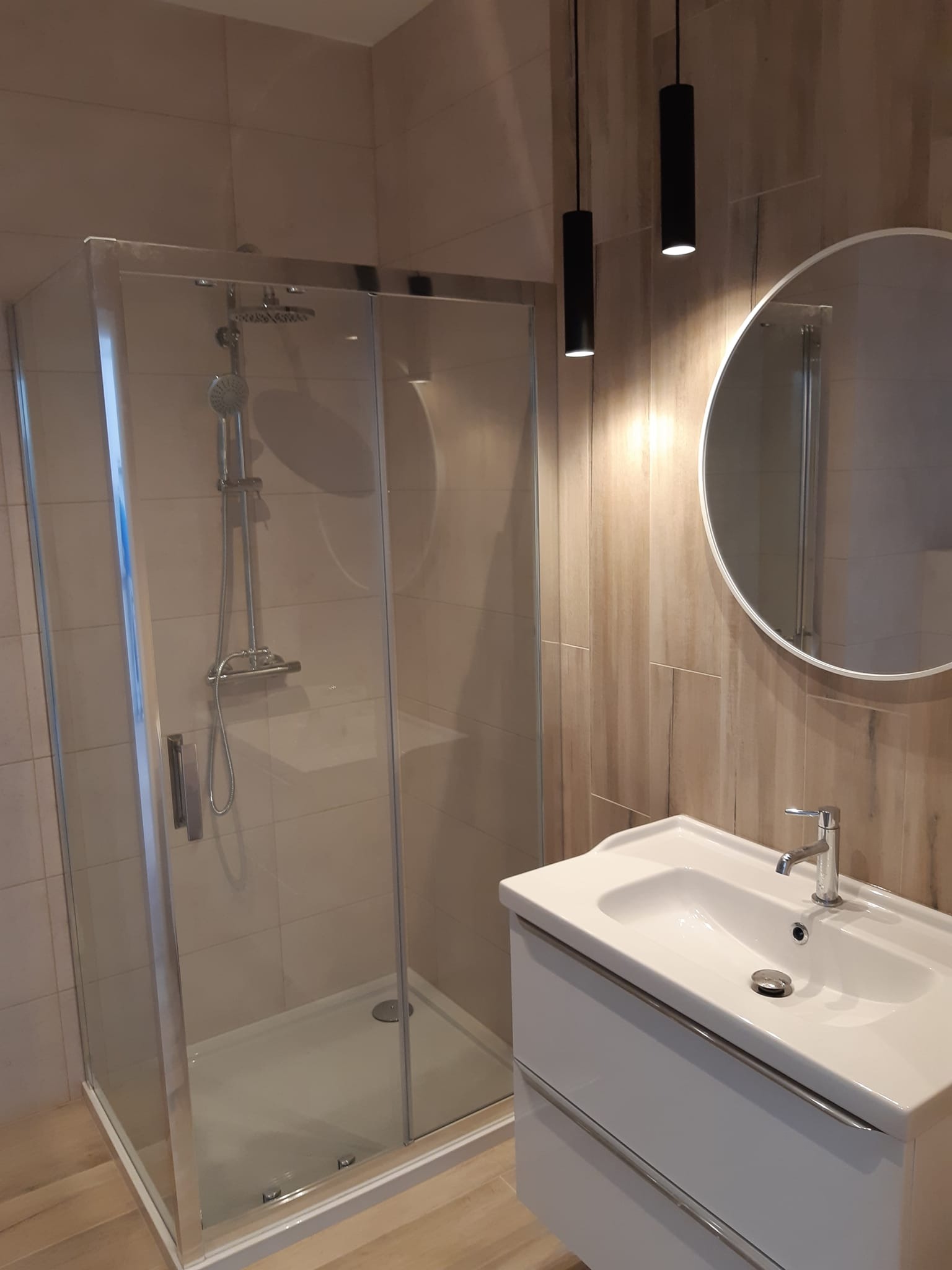 Nowoczesna kabina prysznicowa z chromowaną armaturą i białą umywalką w Keblowie, oświetlona wiszącymi lampami na tle płytek imitujących drewno.