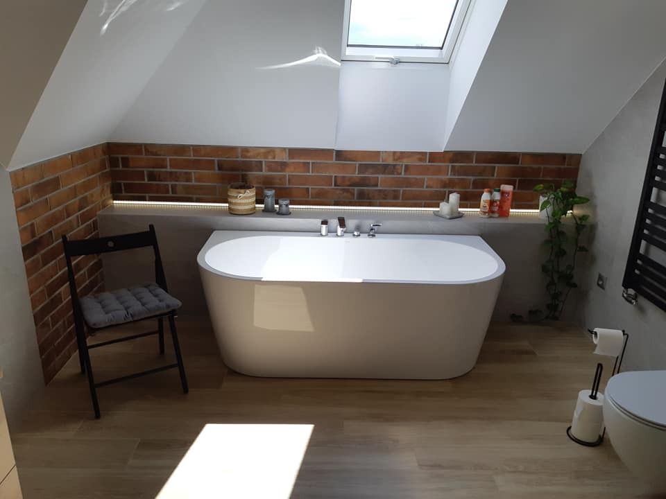 Nowoczesna łazienka na poddaszu z wolnostojącą wanną, ceglaną ścianą i oknem dachowym, uzupełniona o czarne akcenty w postaci krzesła i grzejnika.