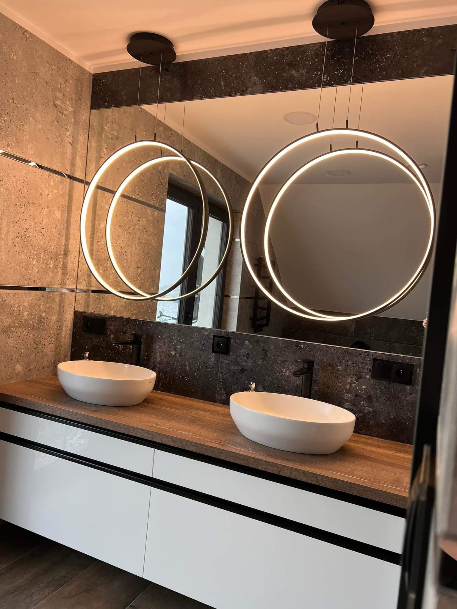 Nowoczesna łazienka z dwoma owalnymi umywalkami nablatowymi, drewnianym blatem, białymi szafkami z czarnymi akcentami oraz okrągłymi lampami wiszącymi z podświetleniem LED.