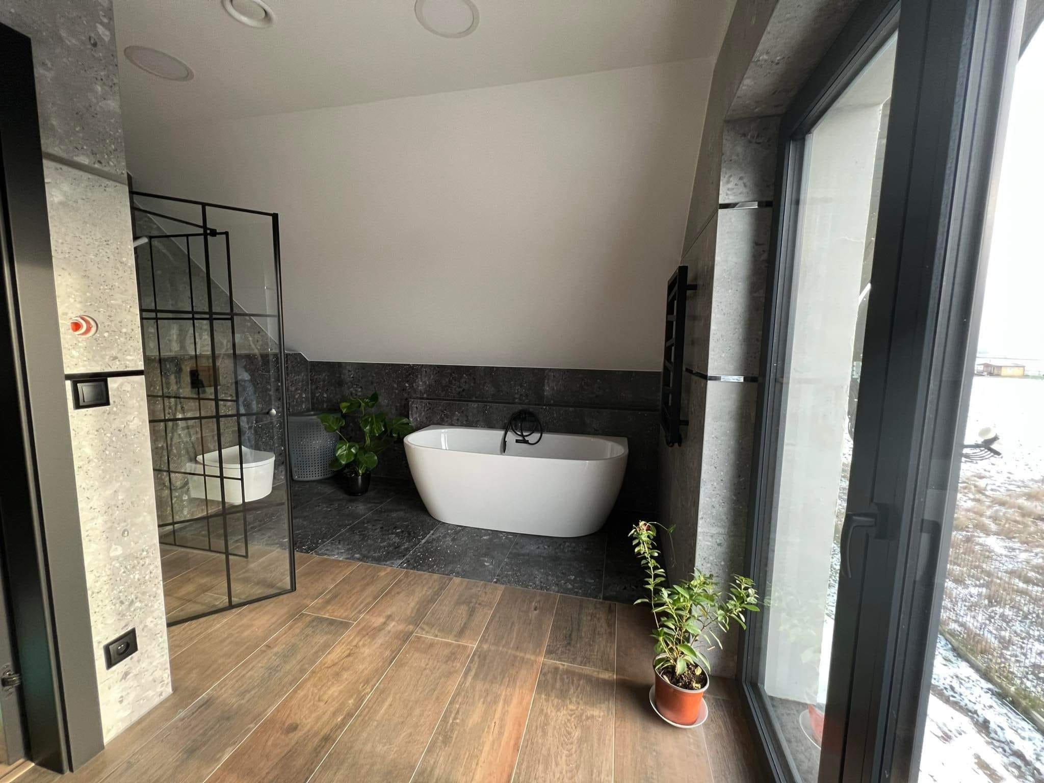 Nowoczesna łazienka na poddaszu z wolnostojącą białą wanną, kabiną prysznicową typu walk-in z czarnymi ramami, drewnopodobną podłogą i roślinami doniczkowymi przy oknie.