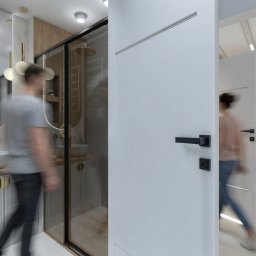 Studio Projektowe Joanna Piekarz - Nowoczesna łazienka z kabiną prysznicową za szklaną ścianą, umywalką na szafce z ryflowanym frontem i okrągłym lustrem. Widoczne osoby w ruchu, białe drzwi z czarną klamką.