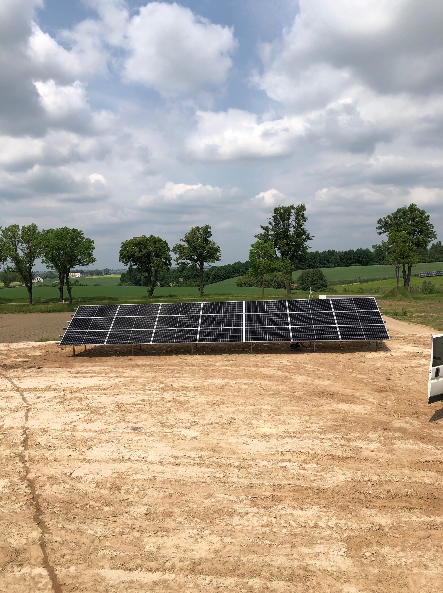 Panel słoneczny na piaszczystym podłożu z widokiem na zielone pola i drzewa pod pochmurnym niebem. Częściowo widoczny biały samochód dostawczy po prawej stronie.