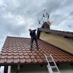 MK Washing - Pracownik w uprzęży czyści dach z brązowej dachówki za pomocą myjki ciśnieniowej. Obok drabina oparta o rynnę. W tle pochmurne niebo i antena.
