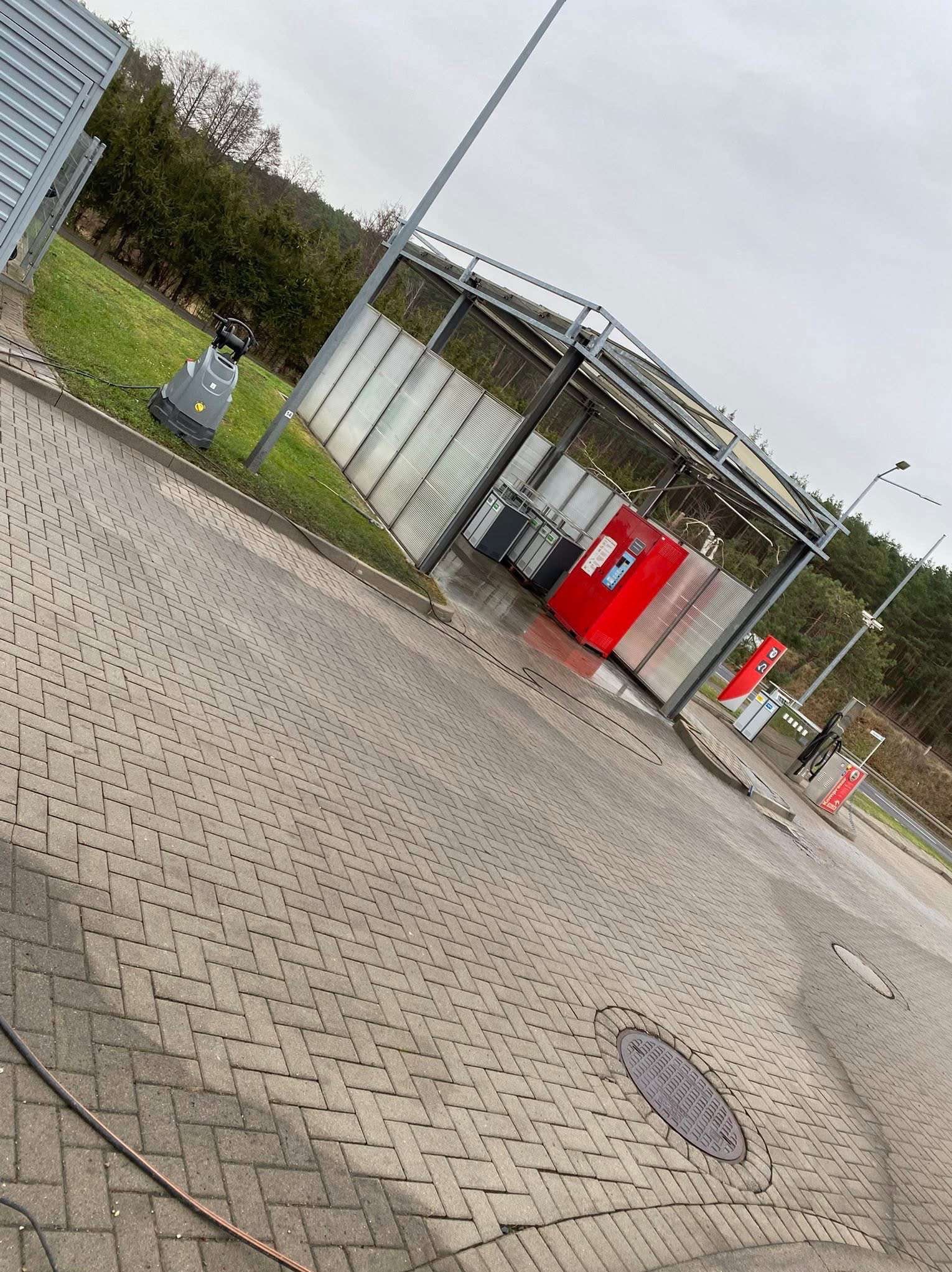 Kostka brukowa po myciu ciśnieniowym, widoczny automat myjący i ślady wody. Ujęcie z perspektywy, lekko pochylone, na tle zieleni i pochmurnego nieba.