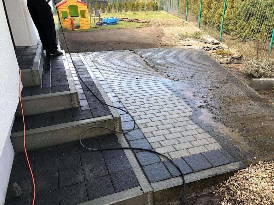 Częściowo umyta kostka brukowa i schody wejściowe z ciemnych płyt, widoczne zabrudzenia i woda. W tle plac zabaw i ogrodzenie.