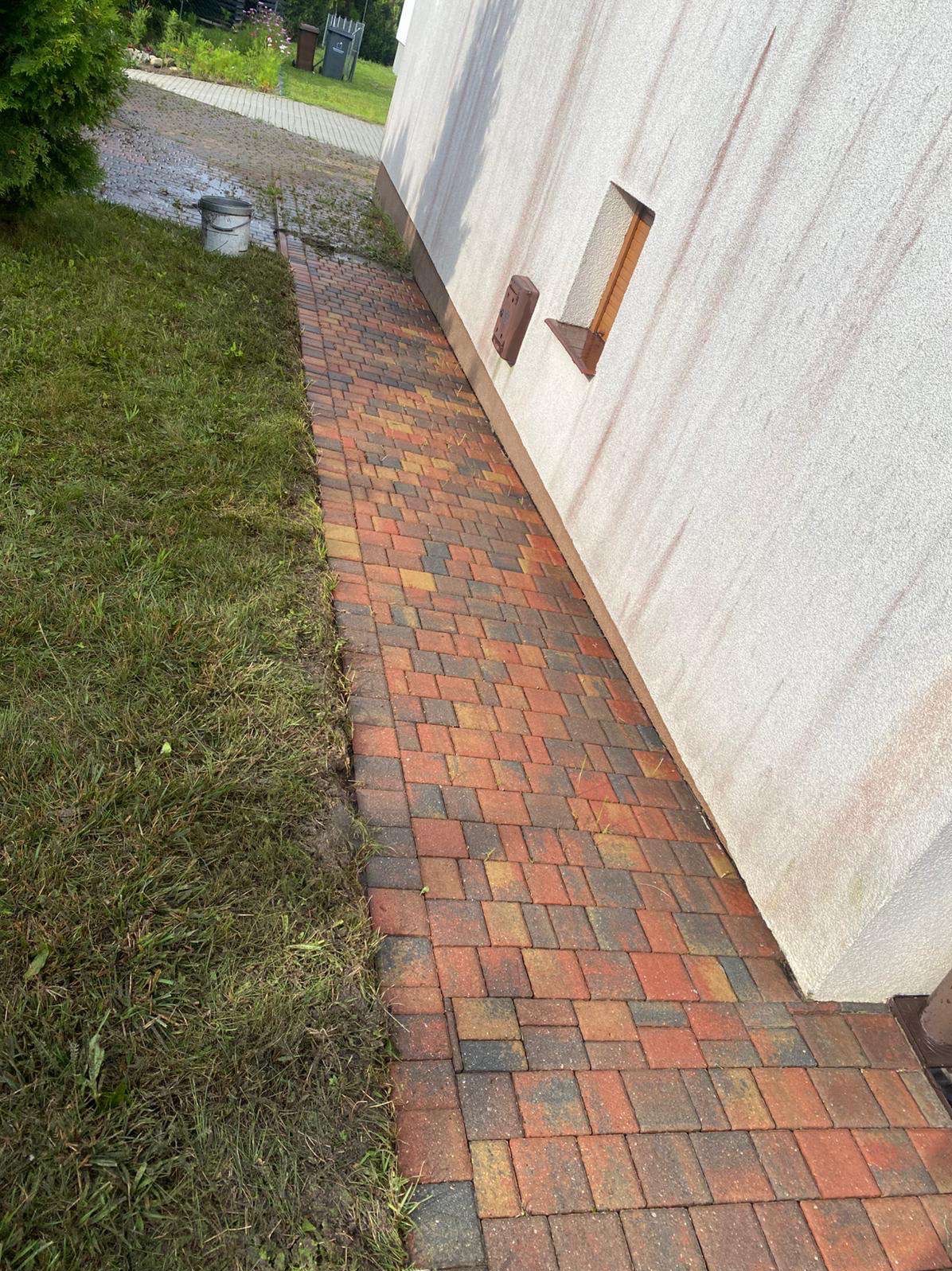 Czerwono-brązowa kostka brukowa ułożona wzdłuż ściany budynku z widocznymi zabrudzeniami i mchem, sugerująca potrzebę czyszczenia. Obok trawnik.